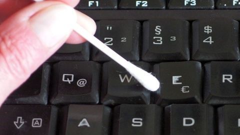 Eine Hand hält ein Ohrenstäbchen, das über die Tasten einer dunklen Tastatur gleitet, um diese zu reinigen.