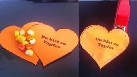 Zwei orangefarbene Herzformen liegen nebeneinander. Auf einem Herz steht "Du bist so tapfer", während das andere mit bunten Süßigkeiten dekoriert ist.