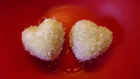 Herzförmige Mandel-Kokospralinen liegen in einer roten Schale und funkeln im Licht. Sie sind mit Kokosflocken überzogen.