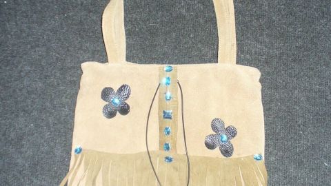 Schlichte Wildledertasche mit fransigen Details und Blumenstickerei hängt vor einem grauen Hintergrund.