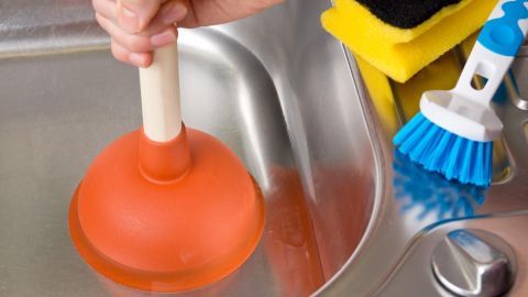 Eine Hand hält einen orangen Pömpel über einem Spülbecken, während Reinigungsutensilien daneben liegen.