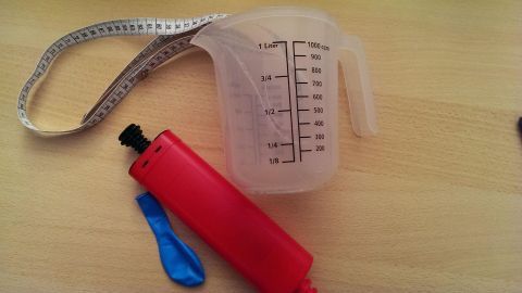 Eine transparente Messbecher steht auf einem Tisch, daneben ein roter Ballon und ein flexibles Maßband, bereit zum Messen.