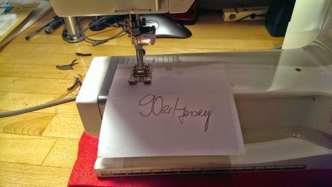 Eine Nähmaschine näht auf einem Tisch ein Blatt mit dem Text "Süßigkeiten". Die Umgebung ist kreativ und funktional.