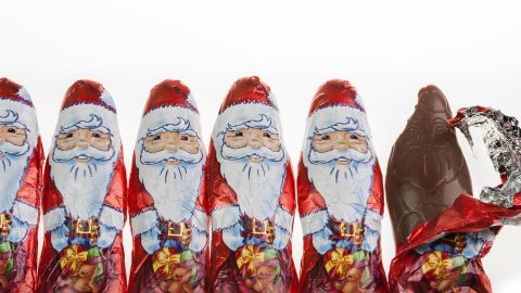 Sechs Schokoweihnachtsmänner stehen in Reihe, während einer bereits geöffnet und teilweise gegessen wurde.
