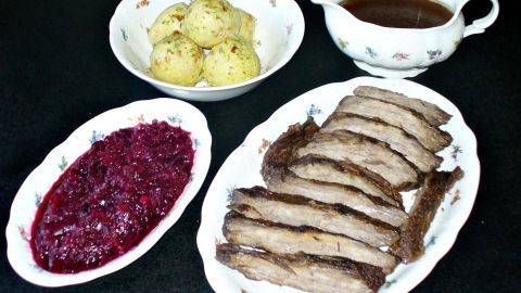 Eingelegter Sauerbraten liegt auf einem Teller, umgeben von Klößen, Soße und Preiselbeermark auf einem festlichen Tisch.