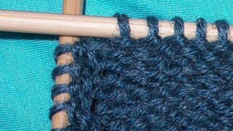 Holz-Nadeln halten einen dunkelblauen Strickfaden in der Nähe einer strahlend grünen Oberfläche, während Runden gestrickt werden.