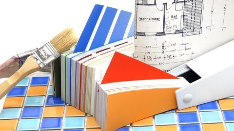 Pinsel liegt auf einem Farbfächer, Fliesenmuster darunter; daneben ein Architektenplan mit Räumen wie Küche und Wohnzimmer.