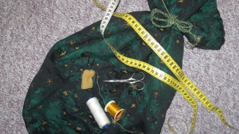 Eine grüne Stofftasche liegt auf einem Teppich und enthält Nähutensilien wie Faden, Nadel und Maßband.