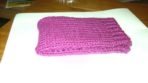 Eine handgestrickte, violette Handytasche liegt flach auf einem weißen Untergrund und zeigt eine einfache, gleichmäßige Struktur.
