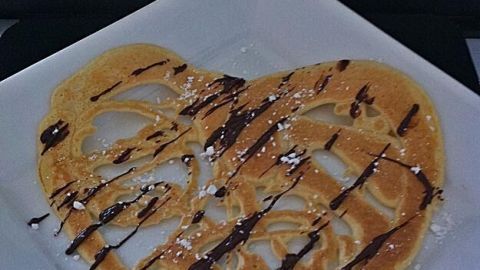 Ein herzförmiger Pfannkuchen auf einem weißen Teller wird mit Schokoladensoße verziert, umgeben von blühenden Blumen.
