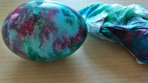 Ein bunt marmoriertes Osterei liegt auf einem Holztisch, verbunden mit einem bunten Stoffstreifen, der Farbe abgibt.