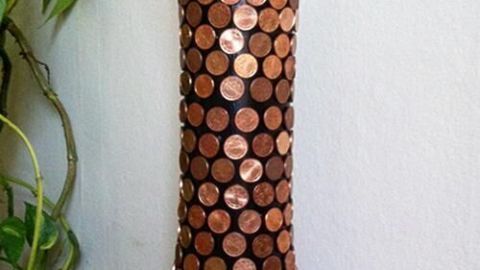 Eine Vase, dekoriert mit 1-Cent-Münzen, steht auf einem Holztisch und hält grüne Pflanzenstängel.