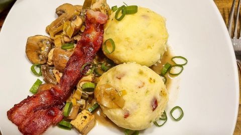 Zwei Kartoffelknödel liegen auf einem weißen Teller, daneben gibt es angebratenen Speck und Pilze in Soße.