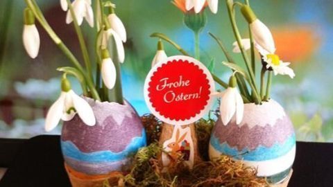 Bunte Eiervasen mit Schneeglöckchen schmücken einen Ostertisch, umgeben von Moos und einer festlichen Botschaft.
