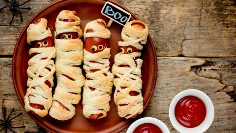 Blätterteig-Würstchen-Mumien liegen auf einem Teller, umgeben von Ketchup und Spinnennetzen, für Halloween dekoriert.