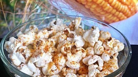Ein Glas würziges Parmesan Popcorn steht im Vordergrund, während frisch geernteter Mais im Hintergrund zu sehen ist.