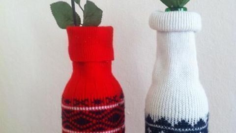 Rote und weiße Flaschen mit Blumen in gestrickten Pullovern stehen dekorativ auf einem Tisch in einem winterlichen Raum.