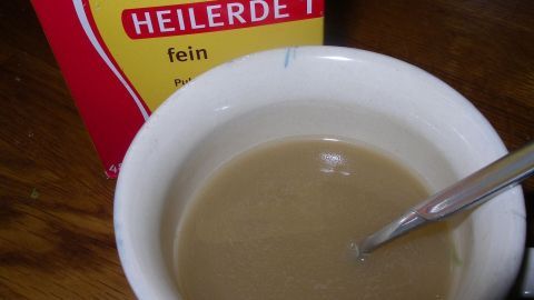 Eine Tasse mit einer braunen Flüssigkeit steht auf einem Tisch, während eine Packung Luvos Heilerde daneben liegt.