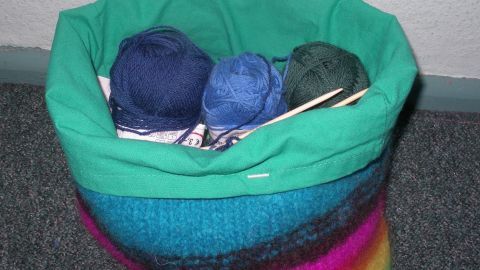 Ein Handarbeitskorb aus Filzwolle steht auf einem Teppich und enthält farbige Wollknäuel und Stricknadeln.