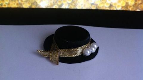 Ein kleines Hütchen liegt auf weißem Papier, dekoriert mit einem goldenen Band und Perlen, vor einem glitzernden Hintergrund.