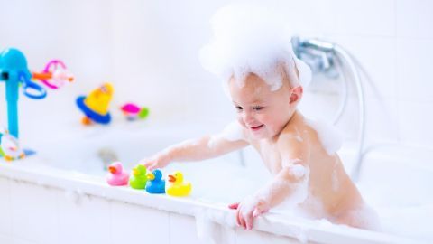 Ein Kleinkind plantscht mit Spielzeugente in der Badewanne, umgeben von bunten Badewannen-Spielzeugen und Schaum.