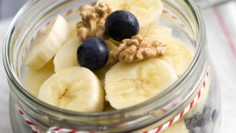 Ein Glas mit Übernacht-Haferflocken zeigt Schichten aus Hafer, Bananen, Blaubeeren und Walnüssen, auf einem hellen Tuch.