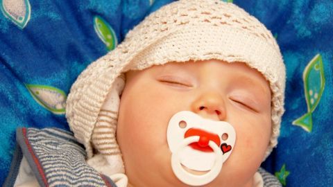 Ein schlafendes Baby mit Schnuller liegt auf einer bunten Decke, die mit Blättern und Wasserzeichen verziert ist.