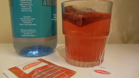 Ein Glas mit roter Brause, darin ein Teebeutel, steht neben einer Wasserflasche. Packungstext: "selam Granatapfel Tee".