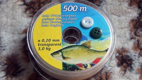 Knöpfe mit Angelschnur annähen - die Angelschnur ist transparent und somit für alle Näharbeiten bestens geeignet. Eine Rolle Angelschnur mit Knöpfen liegt auf einem gemusterten Untergrund, bereit zum Annähen von Knöpfen.