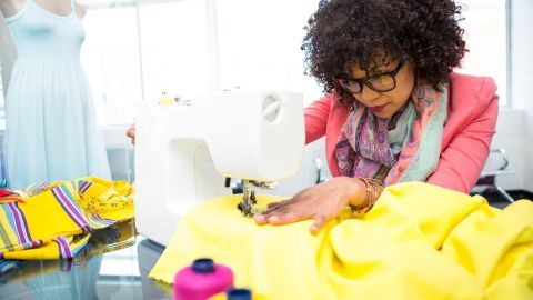 Eine Frau arbeitet konzentriert an einer Nähmaschine, um ein gelbes Kleidungsstück zu nähen, umgeben von Stoffen und Nähutensilien.