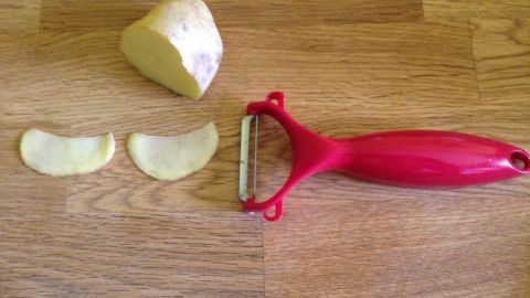 Ein Kartoffelschäler liegt neben einer halben Kartoffel und zwei geschälten Kartoffelscheiben auf einer Holzfläche.