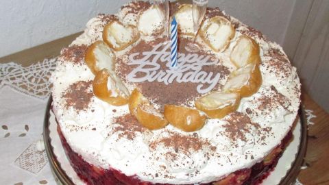 Die Torte steht auf einem Tisch und hat eine Kerze sowie „Happy Birthday“-Schriftzug, umgeben von fruchtigen Windbeuteln.