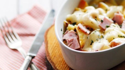Die Kartoffelpfanne mit Schinken liegt in einer weißen Schale, umgeben von rotem Karos-Stoff und einem Holzbrett.