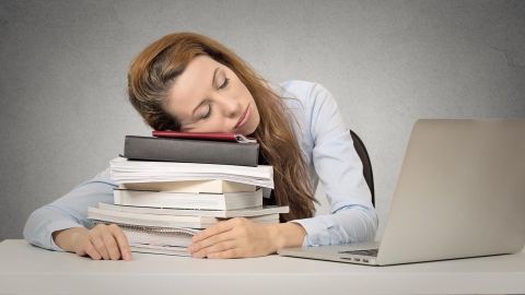 Eine erschöpfte Frau liegt mit dem Kopf auf einem Stapel Bücher, umgeben von einem Laptop, und wirkt geistig erschöpft.