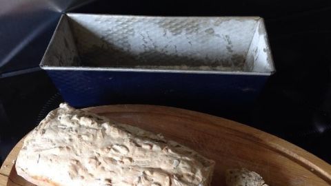 Ein frisch gebackenes Brot ohne harte Kruste liegt auf einem Holzbrett neben einer Backform in einer modernen Küche.