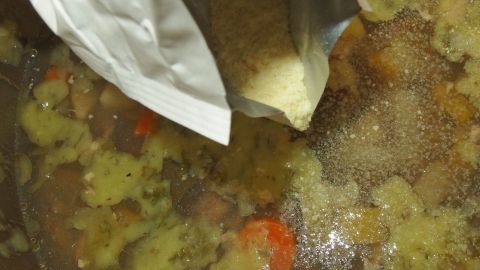 Mit einem einfachen Trick zu dünn gewordene Suppe dickflüssiger machen. Eine Hand gießt eine Pulverpackung in eine große Pfanne mit einer dünnen Suppe, die Gemüse und Fleisch enthält.
