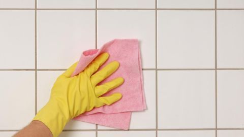 Eine Hand mit einem gelben Handschuh reinigt dunkle Badfugen mit einem pinken Tuch in einem hellen Fliesenbad.