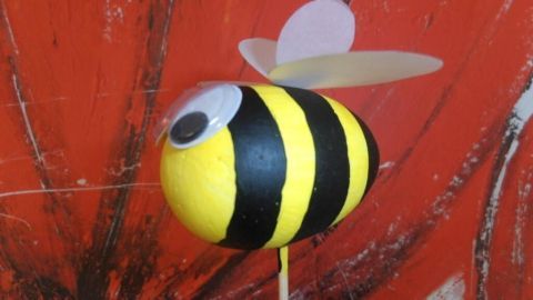 Eine gelb-schwarze Bastel-Hummel mit Flügeln steht auf einem Stock vor einer roten, blumigen Hintergrundgestaltung.