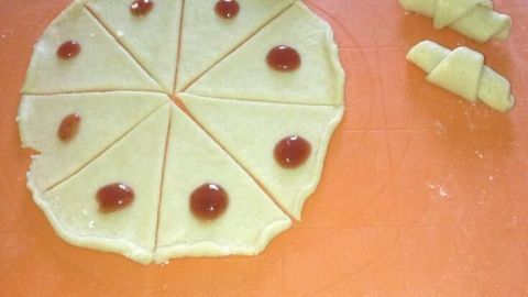 Teigkreise mit Marmeladenklecksen werden zu Rogaliki auf einem orangefarbenen Schneidebrett gerollt.