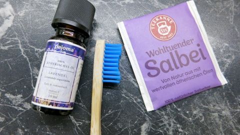 Flasche ätherisches Lavendelöl und Bambuszahnbürste liegen neben Teebeutel „Wohltuender Salbei“ auf einer dunklen Marmorplatte.