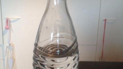 Eine Glasflasche steht auf einem Tisch und enthält Wasser. Der Hintergrund ist hell und schlicht gehalten.