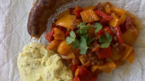 Eine Bratwurst liegt neben einer bunten Gemüsepfanne mit Paprika und einer Portion cremigem Püree auf einem Teller.