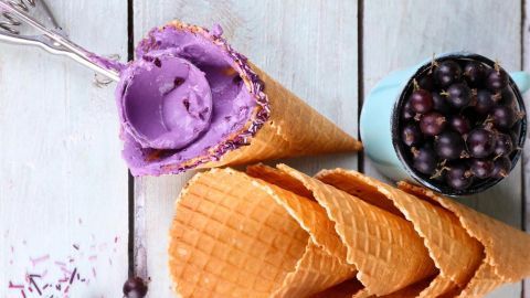 Eiswaffeln liegen auf einem Holztisch, während lila Eiscreme in einer Waffel serviert wird. Runde Schale mit Beeren daneben.