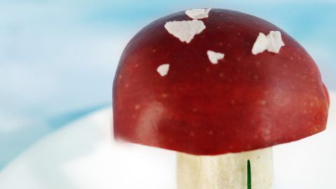 Roter Pilz aus Apfel und Banane steht auf einem weißen Teller, umgeben von frischen Kräutern auf einem hellblauen Hintergrund.