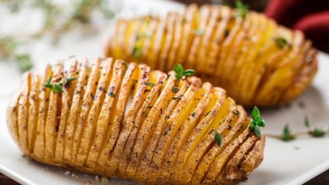 Zwei goldbraun gebackene Hasselbackspotatis liegen auf einem weißen Teller, garniert mit frischen Kräutern und Salz.