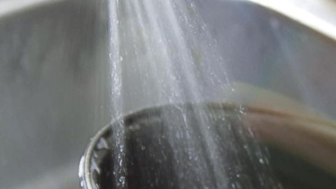 Ein Hand hält einen Topf unter einen Wasserstrahl, um Fett auszuspülen, während die Spüle im Hintergrund sichtbar ist.