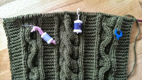 Zöpfe in einem gestrickten, olivgrünen Stück mit farbigen Markierern, die die gestrickten Muster kennzeichnen.