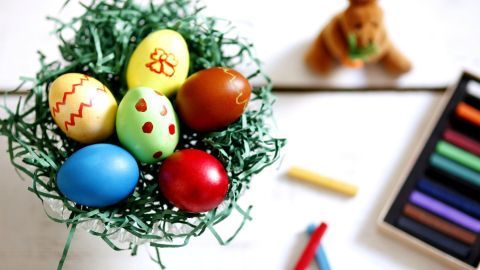 Bunte Ostereier, bemalt mit Wachsmalkreide, liegen in einem grünen Nest aus Gras, umgeben von Zeichenmaterialien.
