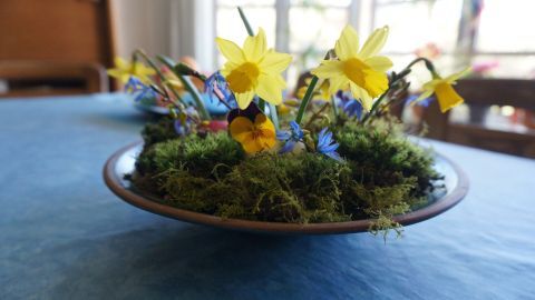 Eine Schale mit Moos und bunten Frühlingsblumen, einschließlich gelber Narzissen, steht auf einem Tisch vor einem Fenster.