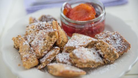 Kaiserschmarrn liegt auf einem Teller, bestäubt mit Puderzucker, daneben ein Glas mit Marmelade in einem hellen Raum.